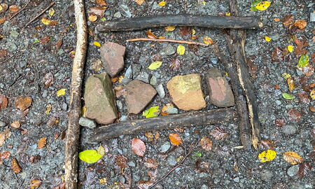 Ein Bild aus Ästen und Steinen auf Waldboden. Die Äste bilden ein Viereck. In der Mitte liegen fünf große Steine.