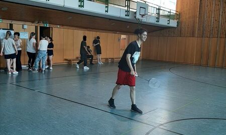 Ein Spieler steht bereit für den Ballwechsel. Er hat einen Badminton Schläger in der Hand. Im Hintergrund stehen einige andere Schülerinnen und Schüler