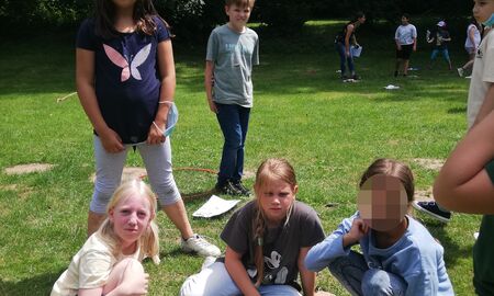 Fünf Schülerinnen und Schüler sitzen und stehen auf einer Wiese um einen auf dem Boden liegenden Reifen, in dem viele Uno-Karten liegen