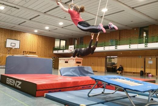 Blick in eine Turnhalle. Im Zentrum sind Matten, Sprungbretter und Kästen aufgebaut. Zwei SchülerInnen schwingen an Ringen über die Matten hinweg.