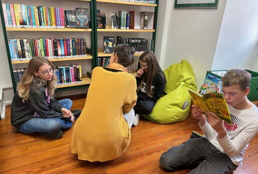 Drei Kinder und eine Erwachsene sitzen bzw. hocken auf dem Boden vor einem Regal voller Bücher. Sie lesen und unterhalten sich.