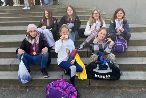 Sieben Mädchen aus der Grundschule sitzen auf einer Treppe und halten Medaillen in die Kamera