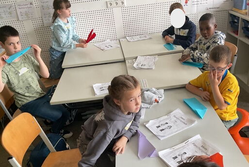 Sechs Kinder sitzen um Tische herum, links im Bild haben zwei Kinder Papierflieger in der Hand, ein Mädchen vorne und drei Kinder rechts im Bild haben Papierflieger vor sich auf dem Tisch liegen