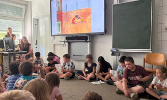 Neun Kinder sitzen nebeneinander auf dem Boden und haben Zettel vor sich liegen. Im Hintergrund sieht man die digitale Tafel mit einem Bild aus einem Bilderbuch. Mit dem Rücken zur Kamera sieht man einige Kinder, die die anderen Kinder anschauen.