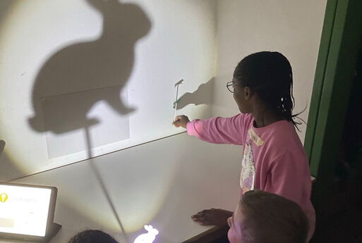 Drei Kinder halten einen Stabpuppenhasen vor ein Licht, ein Stabpuppenhase wirft einen großen Schatten an eine weiße Wand rechts im Bild