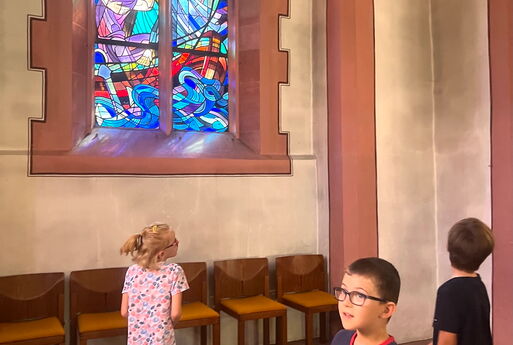 Drei Kinder entdecken die Kirche. In der Mitte des Fotos leuchtet ein buntes Kirchenfenster, auf dem die Geschichte der Sturmstillung abgebildet ist