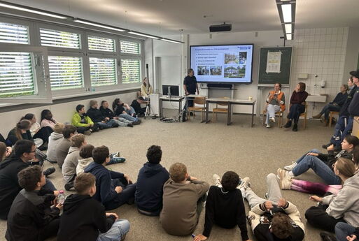 Schüler sitzen im Rund auf dem Boden. Vorne findet ein Vortrag am interaktiven Whiteboard statt.