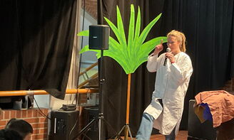 Eine Lehrerin steht auf einer Bühne und hebt ihr rechtes Bein und den rechten Finger nach oben, links neben ihr im Bild sieht man Musikboxen und eine Palme aus Pappe
