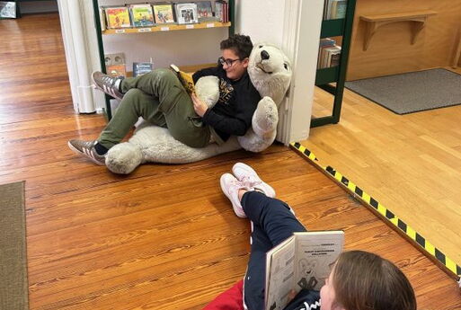 In der Bücherei: Ein Junge hat es sich mit einem Buch auf einem großen Plüscheisbären gemütlich gemacht. Ein Mädchen sitzt auf einem Sitzsack und liest.