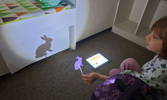 Ein Kind hält einen Stabpuppen-Hasen vor ein Licht und der Hase wirft einen Schatten an eine weiße Wand, auf dem Boden daneben liegt ein iPad
