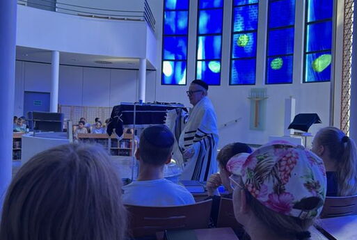 Blick in den großen, in blaues Licht getauchten Innenraum der Synagoge. Vorne sitzen Schüler, in Hintergrund spricht ein Mann.