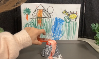 In der Mitte des Bildes sieht man einen Mini-Vulkan, ein Kind schüttet Flüssigkeit hinein, aus dem Vulkan kommt eine schaumige Flüssigkeit raus, im Hintergrund sieht man ein selbst gemaltes Bild