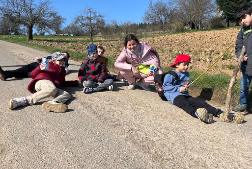 Sechs Jungs und Mädchen aus der dritten Klasse sitzen auf einem Feldweg auf dem Boden, ein Junge steht mit einem Stock rechts im Bild