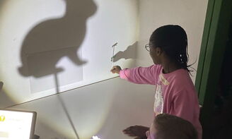 Drei Kinder halten einen Stabpuppenhasen vor ein Licht, ein Stabpuppenhase wirft einen großen Schatten an eine weiße Wand rechts im Bild