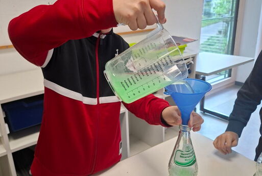 Ein Junge steht an einem Tisch und schüttet mit einem Messbecher eine grüne Flüssigkeit mithilfe eines Trichters in eine Glasflasche