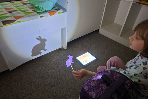 Ein Kind hält einen Stabpuppen-Hasen vor ein Licht und der Hase wirft einen Schatten an eine weiße Wand, auf dem Boden daneben liegt ein iPad