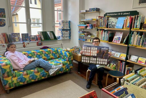In der Bücherei sitzt ein Mädchen auf einem Sofa. Sie hat ein Buch in der Hand und schaut Richtung Betrachter. Ein Junge zeigt ein Plakat.
